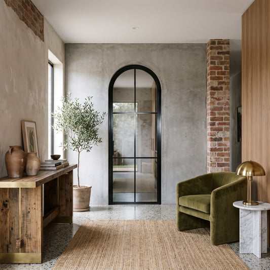 Modern entryway with an arched black steel door, olive tree, velvet green chair, and rustic wood console in a warm, textured home.